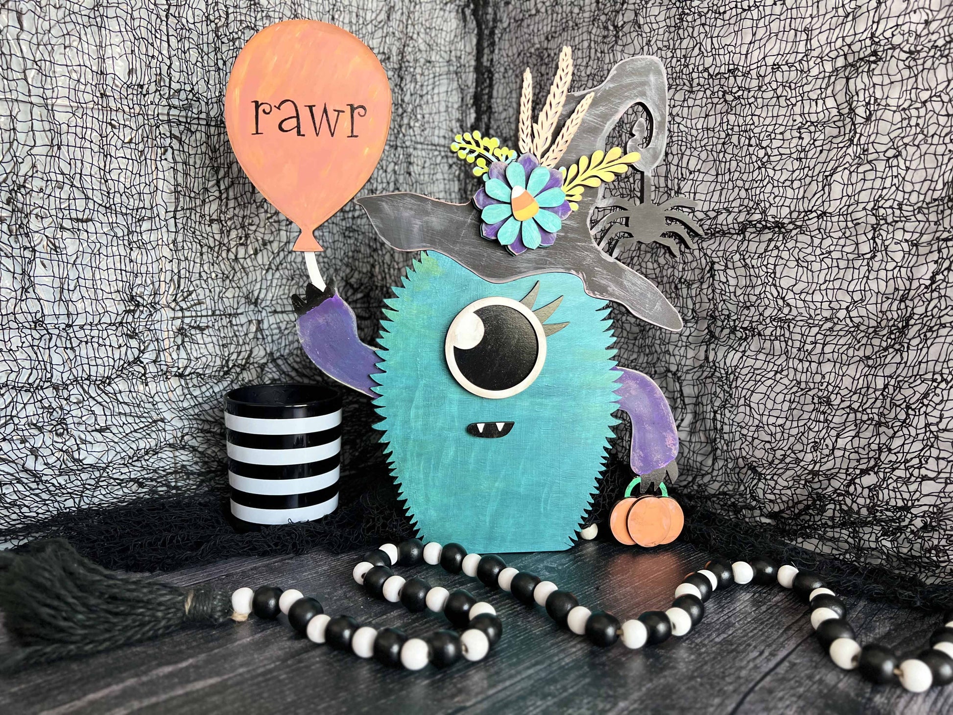 A wooden Halloween turquoise monster in a witch hat holding an orange balloon with the word RAWR printed on it.